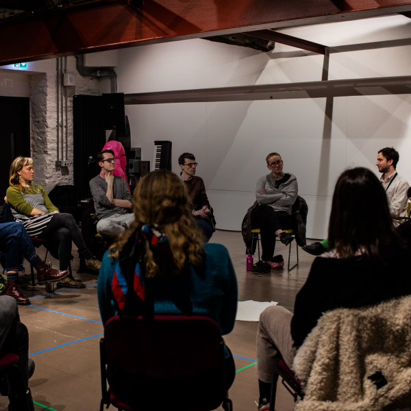 A room of artists sitting on chairs in a large circle, in conversation.