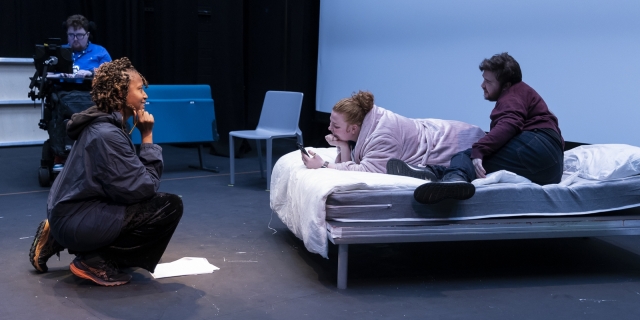 Mid rehearsal, movement director Rudzani Moleya kneels on one knee smiling at two actors on a bed, Georgi Arthur and Nathan Patterson. In the background is director Jamie Hale.