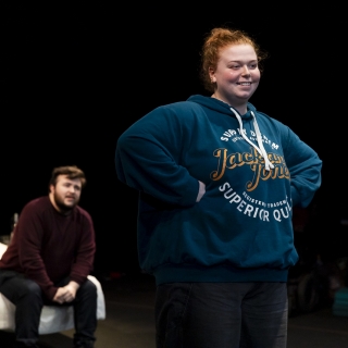 Actors Georgi Arthur and Nathan Patterson performing The Ache of It. Georgi is in focus, smiling at something off camera with hands on hips, whilst Nathan sits behind on the bed, looking on.