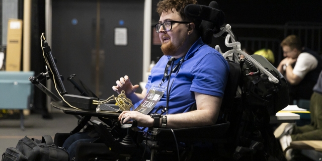 Director, Jamie Hale, in rehearsals. They have red hair and beard and wear a blue shirt with CRIPtic Arts lanyard and sit in their electric wheelchair. They smile with hand raised and mouth open mid sentence.