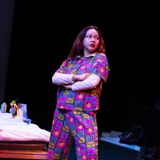 SO SHA performing Pruu the Pidj. She wears a brightly coloured matching shirt and trouser set with pink, blue and green patterns and yellow lemons. She looks to the left with a determined expression on her face with arms crossed and a confident stance.